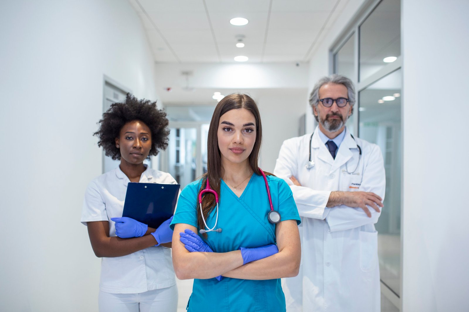 A multi-ethnic group of three doctors and nurses standing in a hospital corridor, wearing scrubs and coats. The team of healthcare workers are staring at the camera and smiling