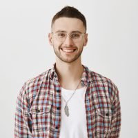 Charming shop assistant ready to offer his help. Portrait of attractive emotive young man with beard in eyewear smiling cheerfully while posing against gray background in studio. Emotions concept.